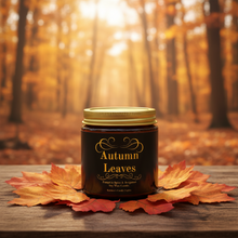 Load image into Gallery viewer, A dark amber glass jar with a gold lid and a label reading 'Autumn Leaves' sits on a wooden surface surrounded by orange and red maple leaves, with a blurred autumn forest background.