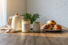 Load image into Gallery viewer, Two glass jar candles labeled 'Cinnamon Buns' by Karmen’s Tearoom , one large and one small, both with gold lids and white wax, set on a cork-like surface with a cardboard backing behind them.