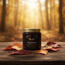Load image into Gallery viewer, A dark amber glass jar candle with a gold lid and a label reading 'Hello Autumn' sits on a rustic wooden surface scattered with red and orange autumn leaves, with a blurred golden forest background.